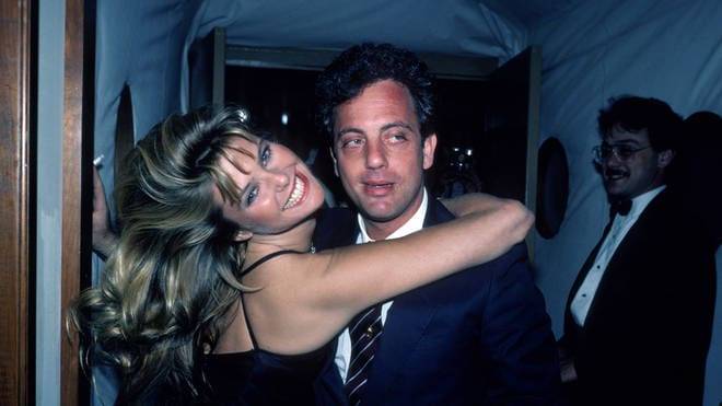 Billy Joel posing with a woman at an event, both smiling and dressed formally.