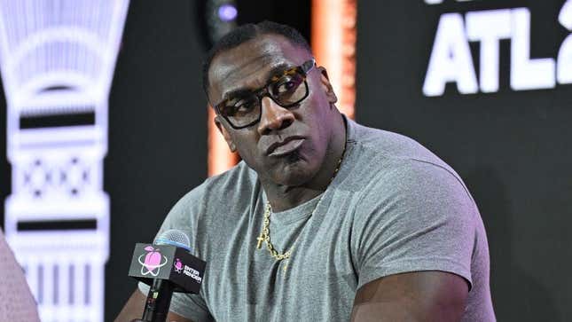 Shannon Sharpe wearing a gray shirt and glasses speaking at a press conference with a microphone in front of him.
