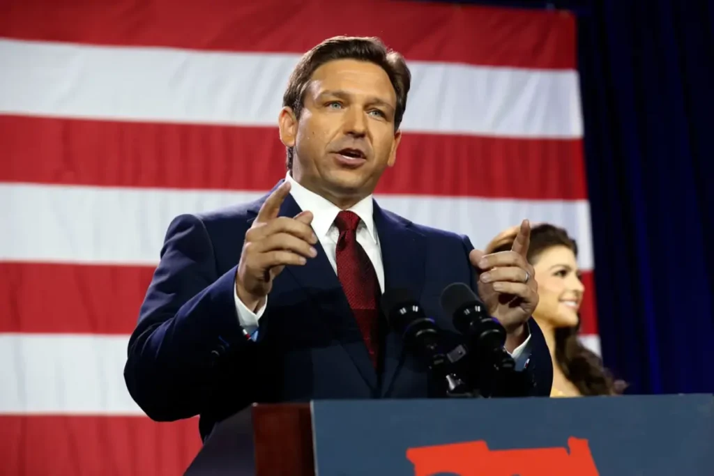 Ron DeSantis speaking at a podium with a microphone, American flag backdrop, and a woman standing beside him.