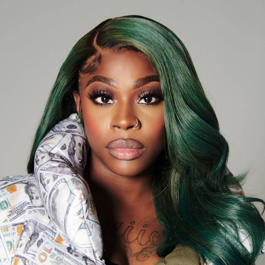 Close-up portrait of Juiicy 2xs with long green hair and a patterned jacket featuring dollar bills.