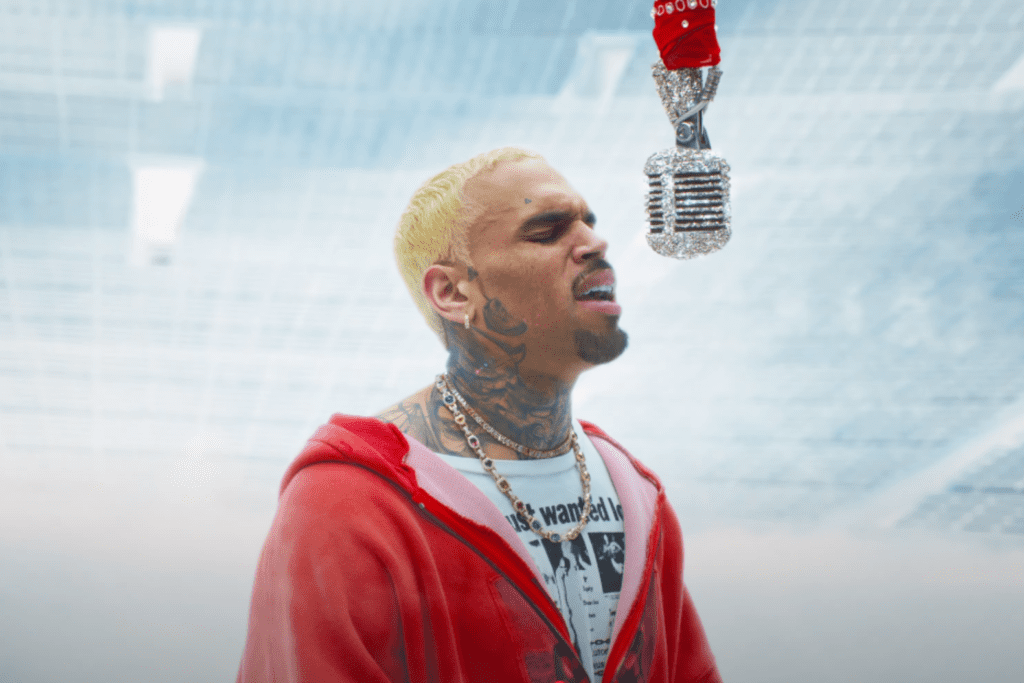 A man with blonde hair and tattoos singing into a vintage microphone while wearing a red hoodie.