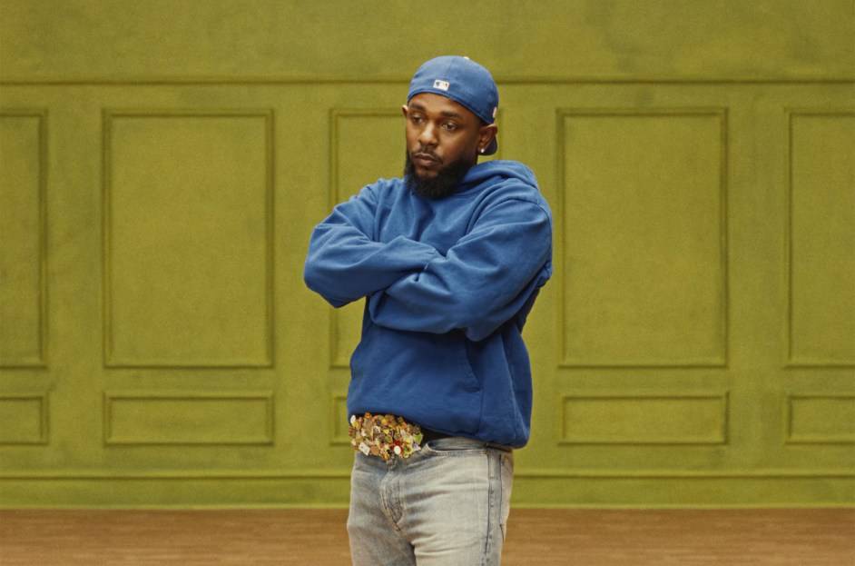 Kendrick Lamar wearing a blue hoodie and jeans, standing with arms crossed in front of a green wall.
