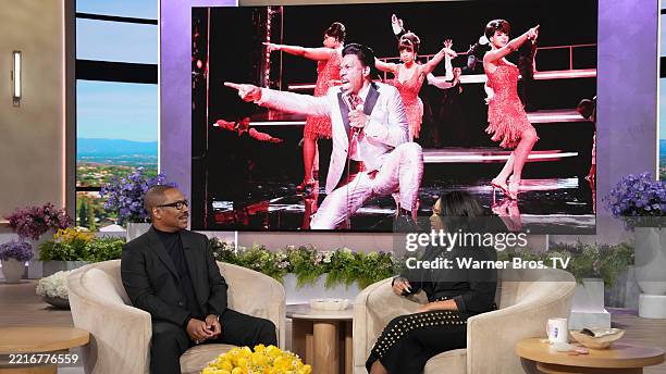 Two guests seated on a talk show set with a large screen displaying performers in the background.