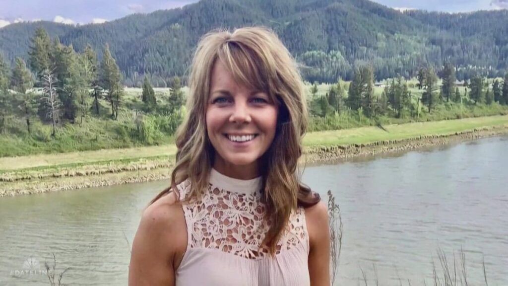 A woman with long brown hair smiling near a river surrounded by trees and mountains.
