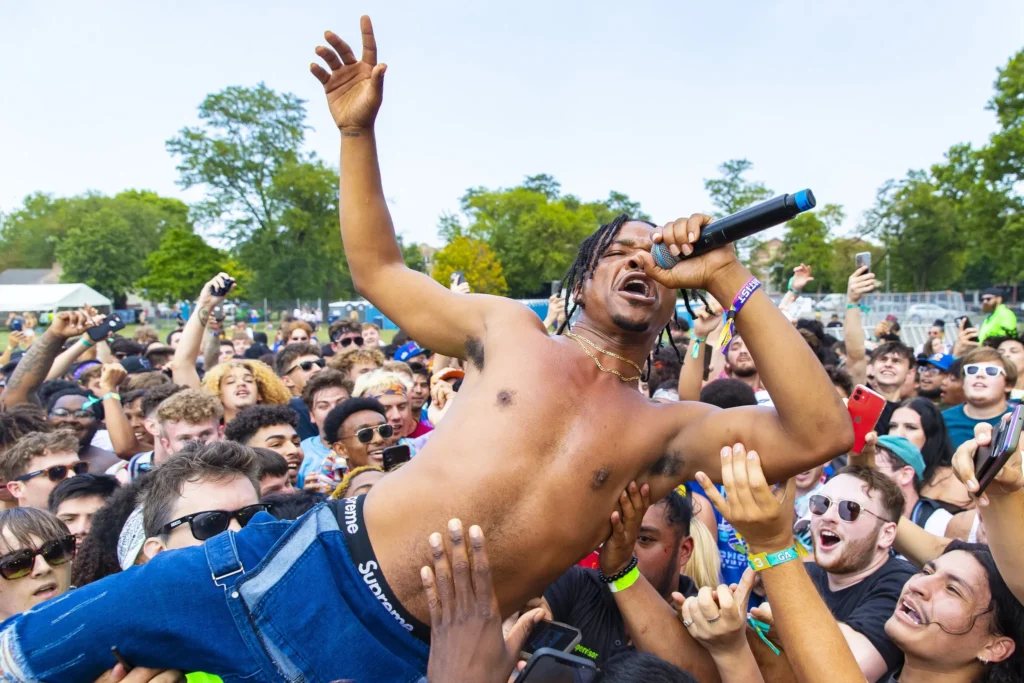Male performer with a microphone singing while being supported by a crowd of fans at an outdoor event.