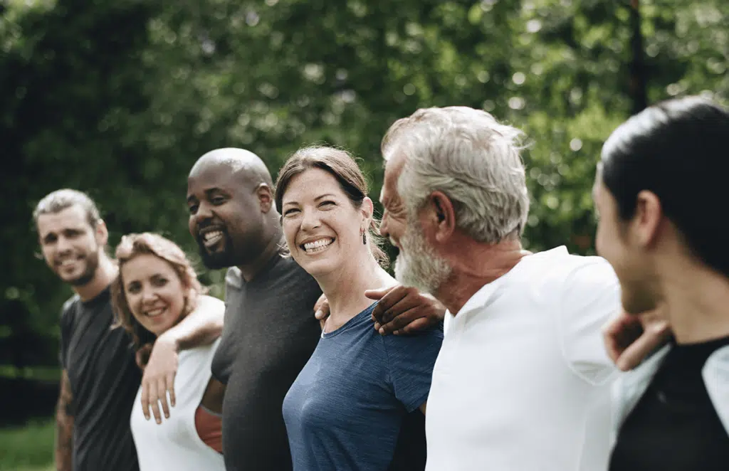 A diverse group of six people smiling and standing in a line outdoors with arms around each other.