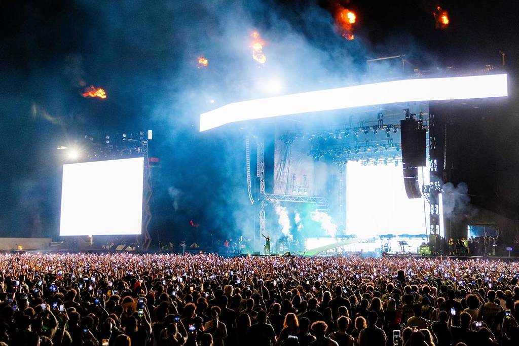 Crowd of concert attendees holding up phones in front of a large stage with bright lights and smoke effects.