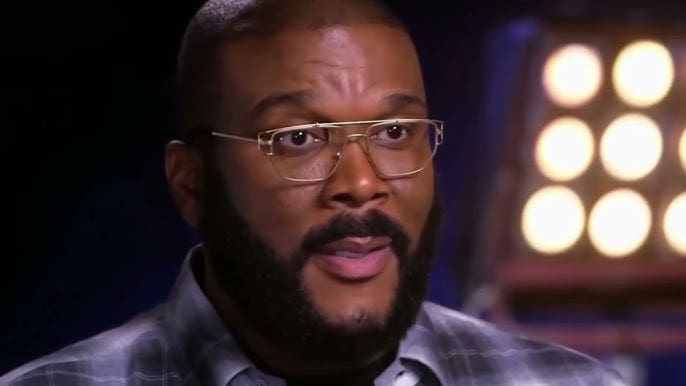 A man wearing glasses and a plaid shirt, with a beard, speaking in an interview setting.
