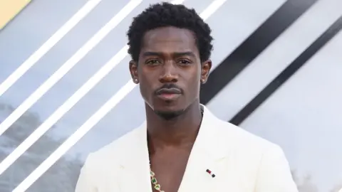 A man wearing a white suit with short curly hair and a patterned shirt underneath.