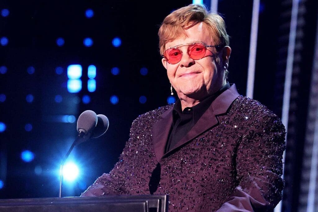 Elton John wearing sunglasses and a sequined jacket while standing at a podium with microphones.