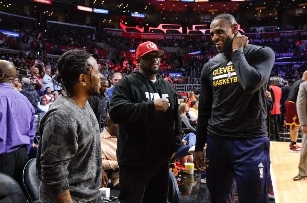 LeBron James and Kendrick Lamar talking at a basketball game with a crowd in the background.