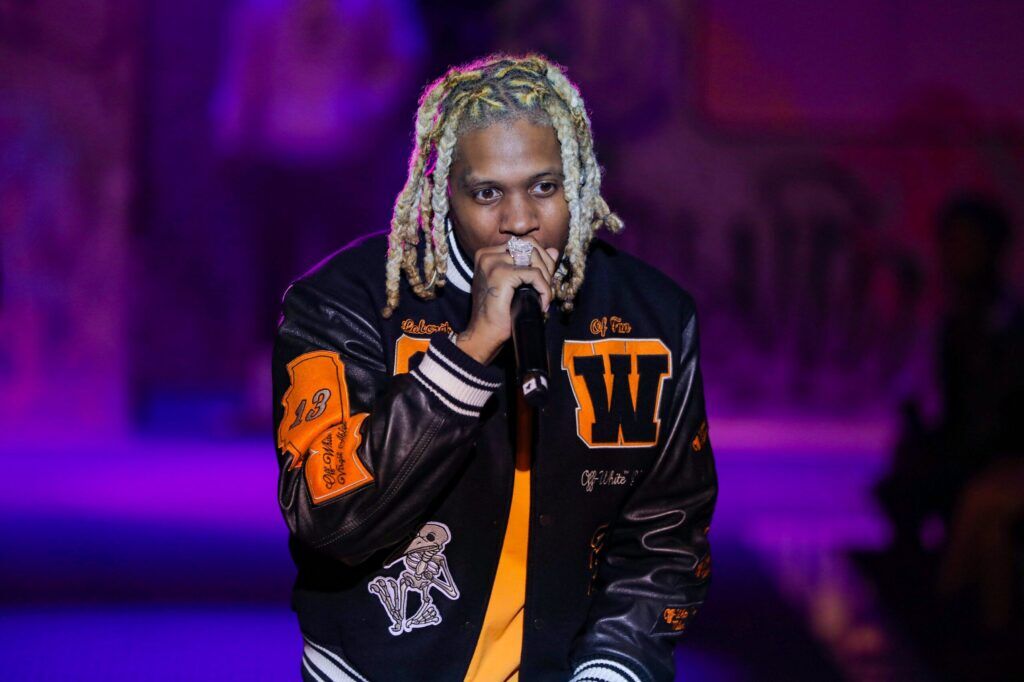 Lil Durk wearing a black and orange jacket while performing with a microphone on stage.