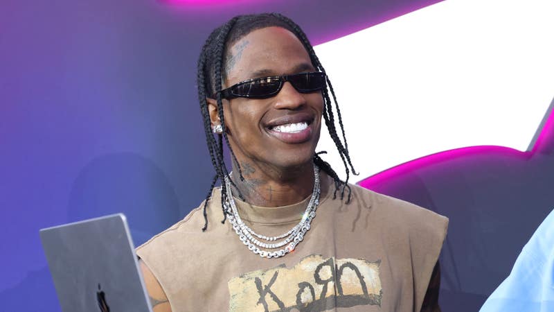 Travis Scott smiling while holding a tablet at an event, wearing sunglasses and a casual outfit.