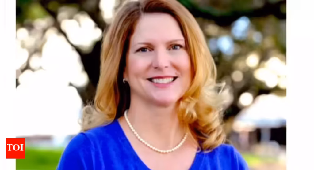 A woman with long hair wearing a blue top and pearl necklace, smiling at the camera.