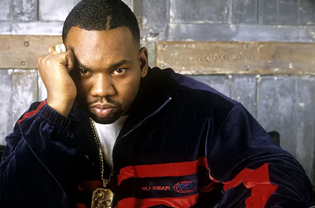Portrait of Raekwon, a hip-hop artist, wearing a navy and red tracksuit with a gold chain.