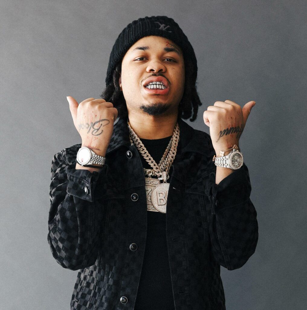 Man wearing a black patterned jacket and multiple silver necklaces, showing a thumbs-up gesture.