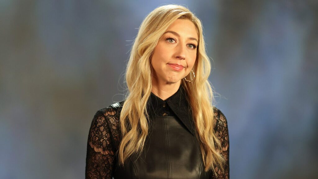 A woman with long blonde hair wearing a black outfit, looking slightly to the side with a neutral expression.