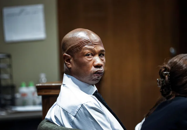 A man with a bald head wearing a white shirt and black tie, seated and looking towards the camera.
