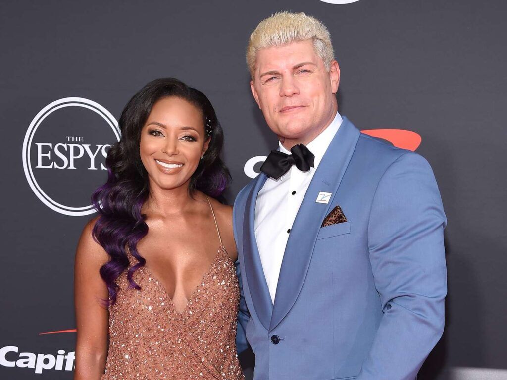 This photo shows Cody Rhodes and Brandi Rhodes dressed elegantly on the red carpet at the ESPYS. Cody is wearing a sharp light blue tuxedo with a black bow tie and white shirt, while Brandi stuns in a sparkling rose gold gown with a deep neckline, her wavy hair styled perfectly. The iconic ESPYS logo is visible in the background, highlighting the event’s prestigious atmosphere.