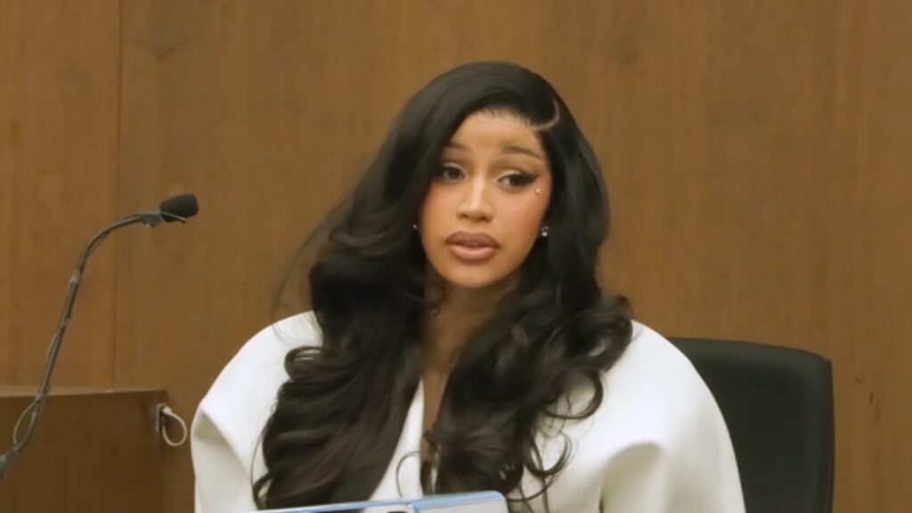 Cardi B seated at a courtroom table while testifying, wearing a white outfit with long black hair.