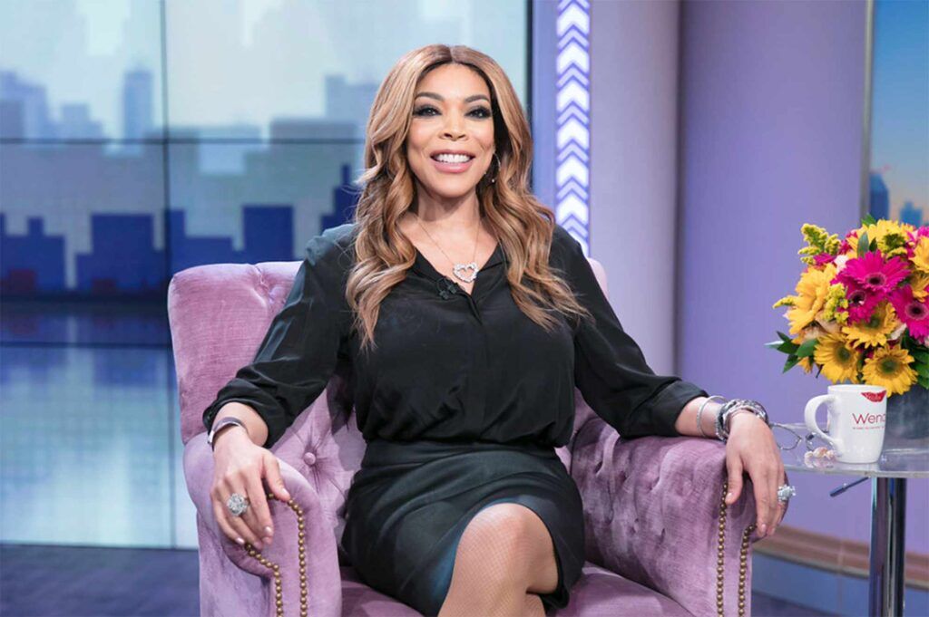 Wendy Williams with long hair sitting on a purple chair, smiling at the camera.