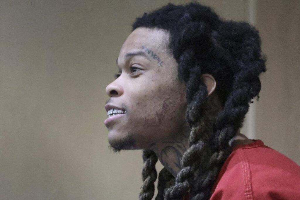 Profile view of an individual with braided hair and facial tattoos, wearing a red outfit.