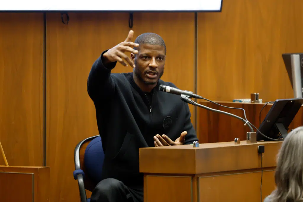 A man speaking at a microphone during a formal hearing, gesturing with his hands.