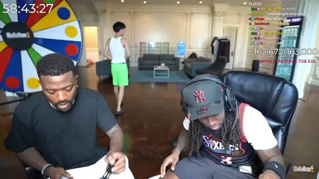 Ray J and Kai Cenat sitting at a table with a colorful wheel in the background and a person walking in the distance.