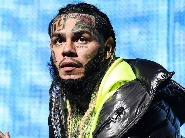 Tekashi wearing a black puffer jacket with colorful tattoos visible on his face during a performance.