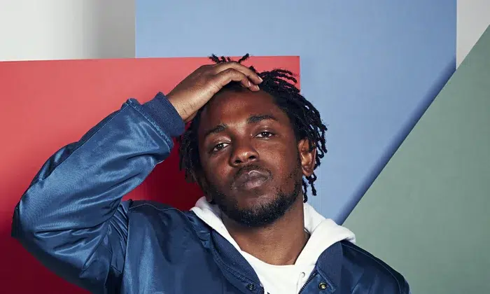 Kendrick Lamar posing with a hand on his head against a colorful backdrop.