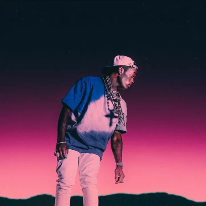 Lil Uzi Vert wearing a blue and white shirt with a cross necklace, standing against a gradient pink and purple background. 4M.