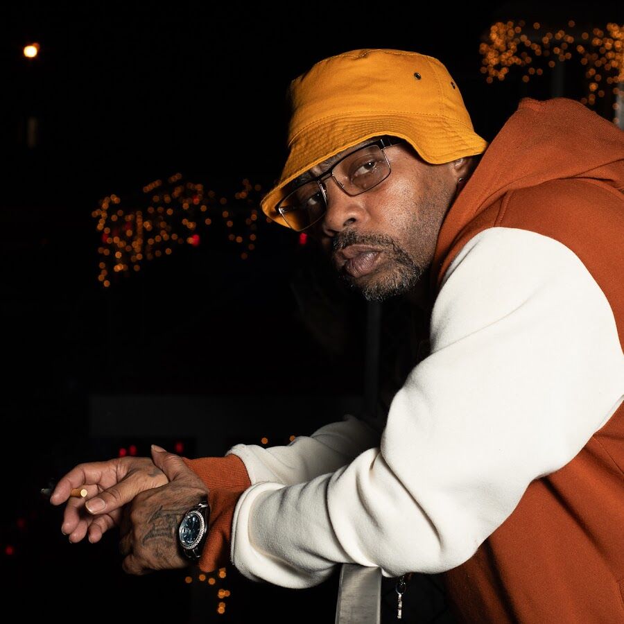 Man wearing an orange bucket hat and brown hoodie, leaning against a railing at night.